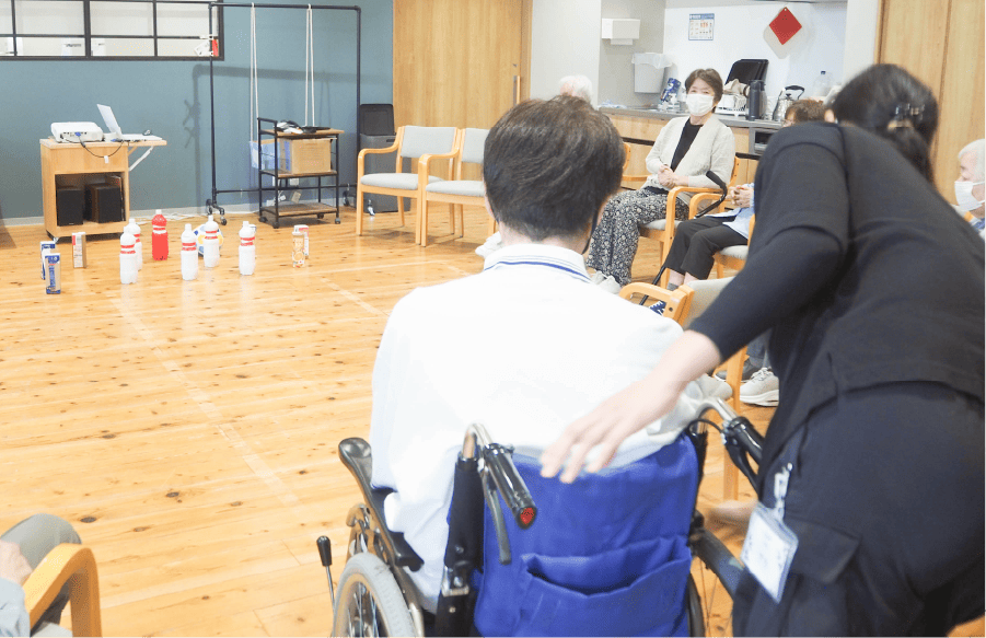ケアマネージャーとの連携も密に行い、必要な支援を行います - 神戸市垂水区・塩屋のデイサービスセンター倖糸(こいと)で床に並べられたペットボトルでボーリングに挑戦中の車椅子の男性と、補助に寄り添っているスタッフの様子